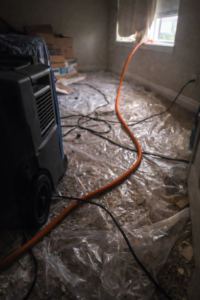 Dehumidifier and drainage hose running on plastic sheeting with cords and debris in a water damaged bedroom during drying process in Layton Utah