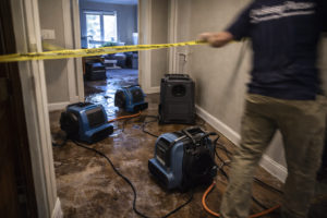 Water damage restoration equipment including air movers and dehumidifier being set up in a flooded residential hallway with visible water and technician in motion in Layton Utah