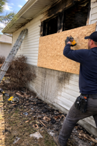 Technician securing plywood over broken fire damaged window with ladder and debris visible outside a residential home in Layton Utah