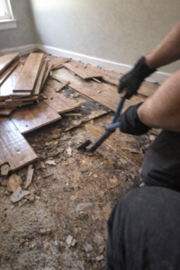 Technician removing warped hardwood flooring with pry bar exposing damaged subfloor and debris during water damage cleanup in Layton Utah home
