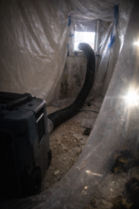 HEPA air scrubber running inside plastic containment with exhaust hose to window and mold damaged walls in background during remediation in Layton Utah