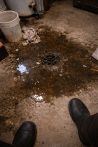 Dark contaminated water spreading across basement floor from floor drain with debris and partially visible rubber boots during sewage backup in Layton Utah
