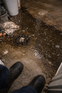 Sewage backup spreading from floor drain across concrete basement floor with debris and technician boots visible during emergency cleanup in Layton Utah