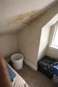 Water leaking from stained and bubbling ceiling near roofline with bucket catching drips in a residential Layton Utah home