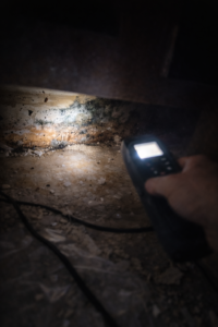 Flashlight revealing moisture damage and early mold growth under kitchen cabinet with debris and technician hand holding tool in Layton Utah home